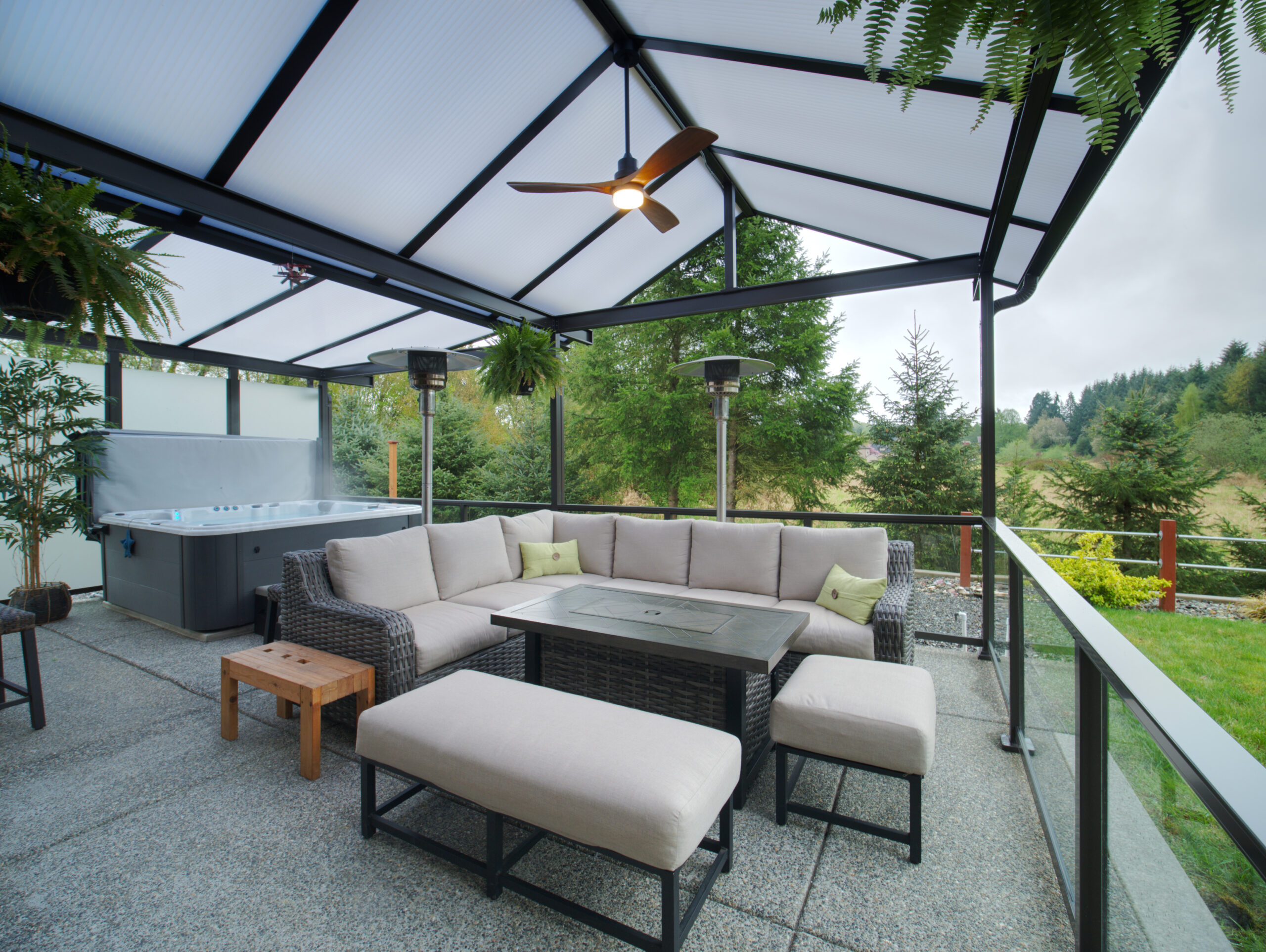 Custom covered patio with double-wall acrylic roof, outdoor sectional seating, heaters, ceiling fan, and hot tub overlooking a landscaped Pacific Northwest backyard.
